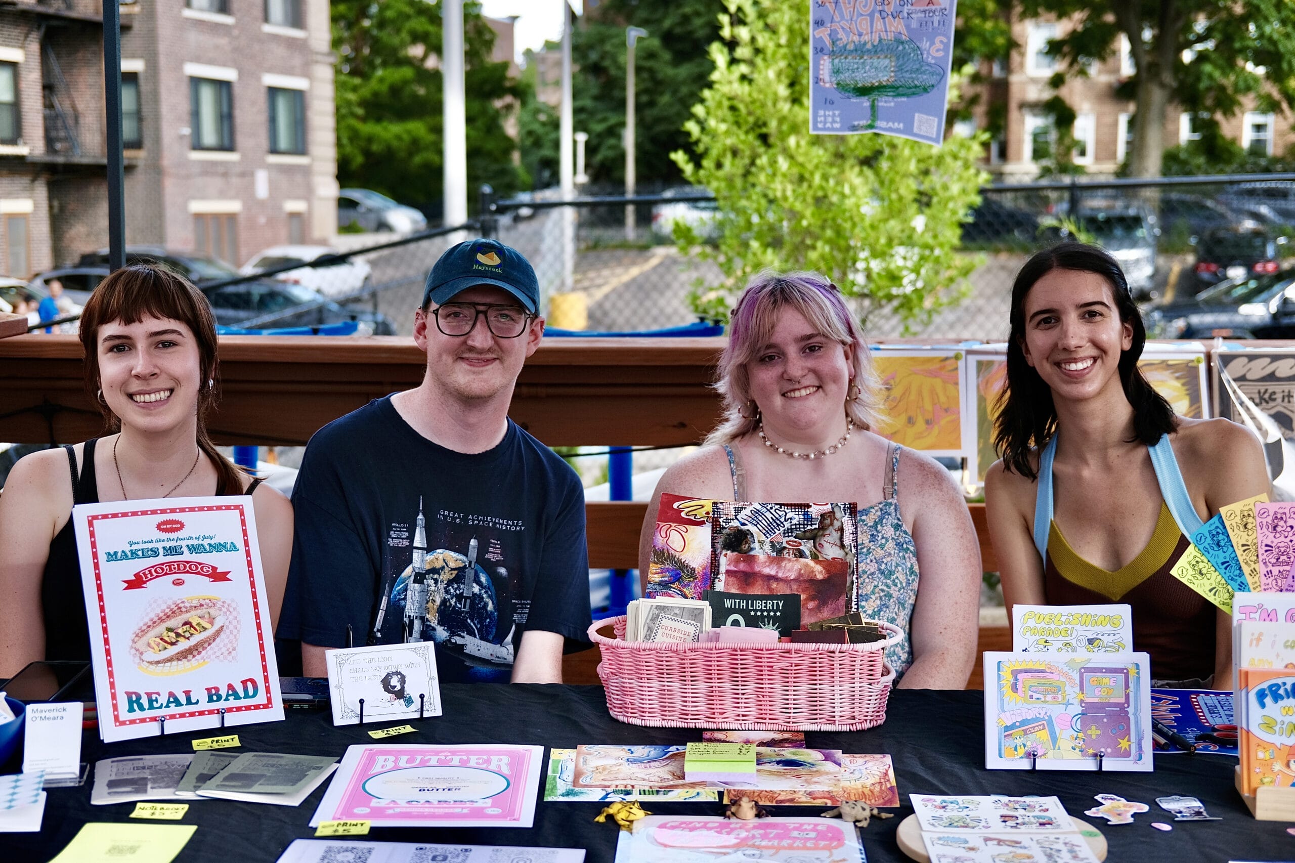 Four people are seated at a table, exuding a chill vibe as they display various zines, stickers, and colorful artwork. They smile warmly amid the urban outdoor scenery of Mark Night Market, with buildings and trees framing the lively backdrop.