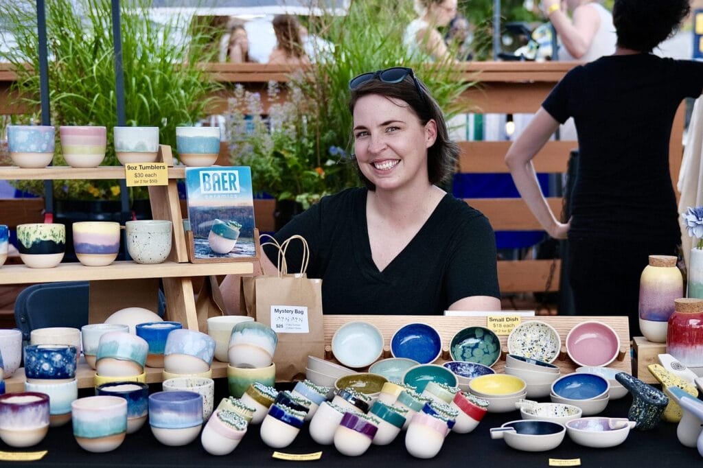 A smiling person sits at a booth at the night market, displaying colorful handmade pottery, including bowls and cups. Signs with prices adorn the table while the person, exuding a chill vibe in black with sunglasses on their head, is framed by lush green plants in the background.