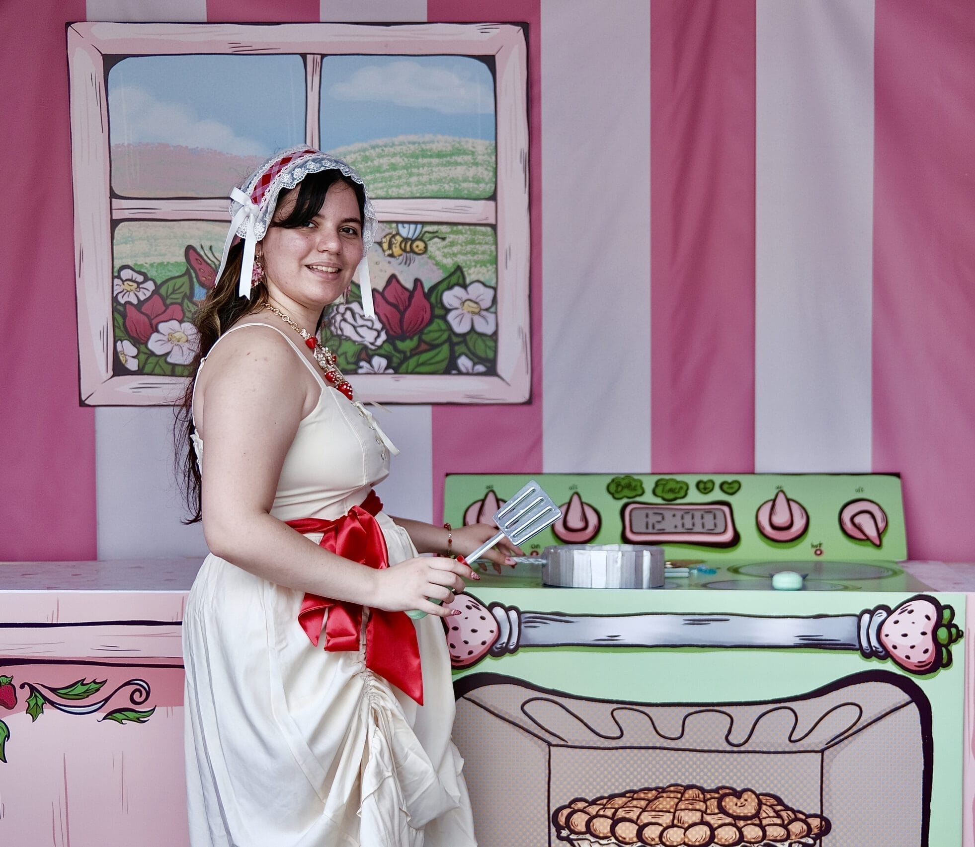 In a decorative kitchen set, a person in vintage-style attire with a bonnet and apron stands artfully holding a spatula. They smile at the camera against pink stripes and whimsical stove illustrations, exuding an effortlessly chill vibe.
