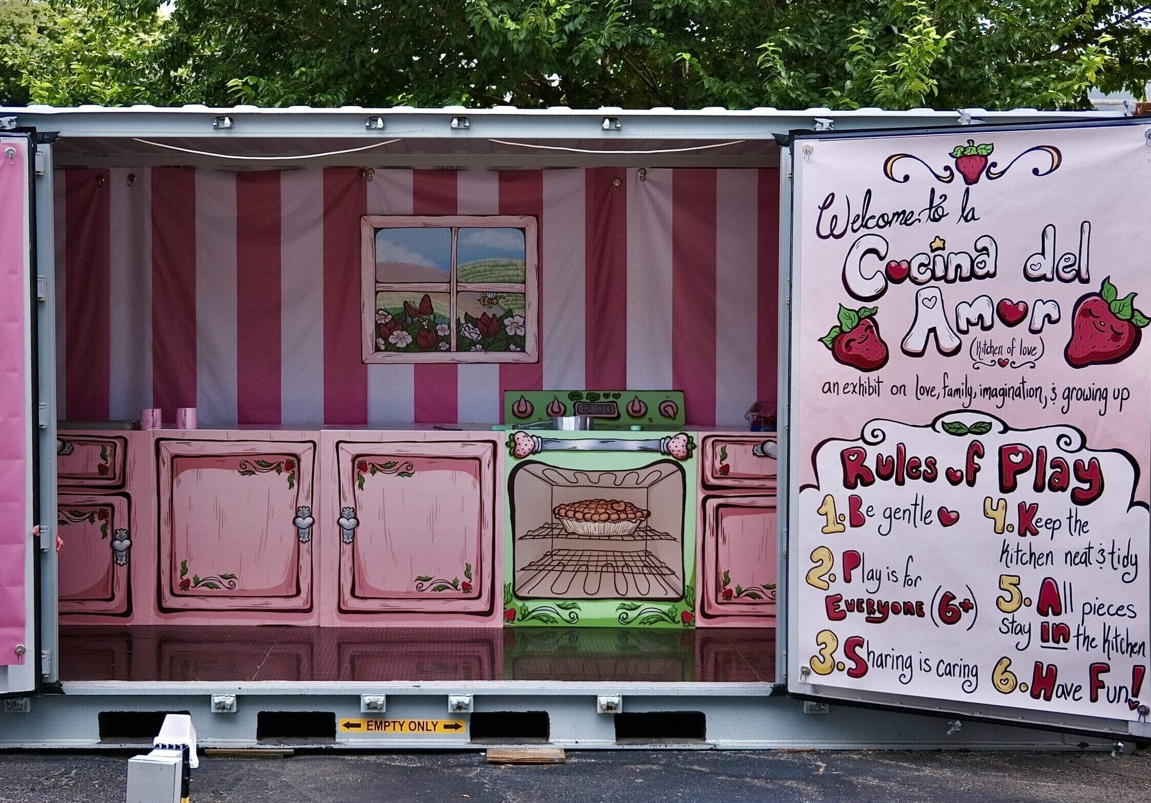 A whimsical kitchen setup inside a container, painted with pink cabinets and strawberries. A sign reads Cocina del Amor alongside playful rules like Be gentle and Have Fun. This colorful, inviting scene exudes an artful, chill vibe amid the trees, perfect for a night market atmosphere.