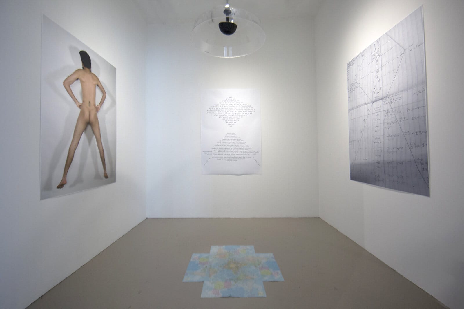 The gallery room features artwork including a large photo of a person from behind on the left wall, a handwritten text piece at the center, and a geometric design on the right. A map-like creation adorns the floor beneath the watchful ceiling surveillance dome, echoing themes from the MassArt Summer Lecture Series.