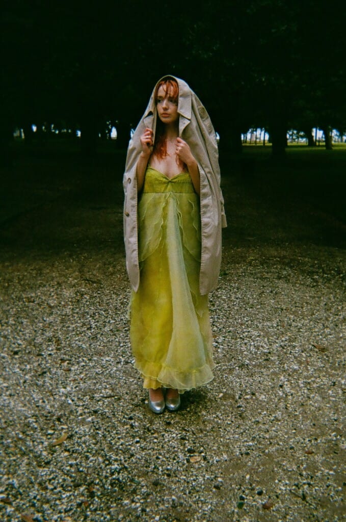 A woman standing in the grass in a green silk dress with a coat over her head.