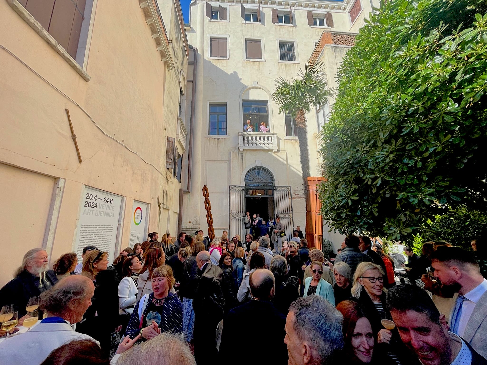 A large crowd gathers in a courtyard outside a historic, light-colored building. Among the chatter, someone admires intricate Venetian Glass. The building has tall windows and a balcony with a few people standing on it, possibly including James McLeod. A tree shades part of the area.