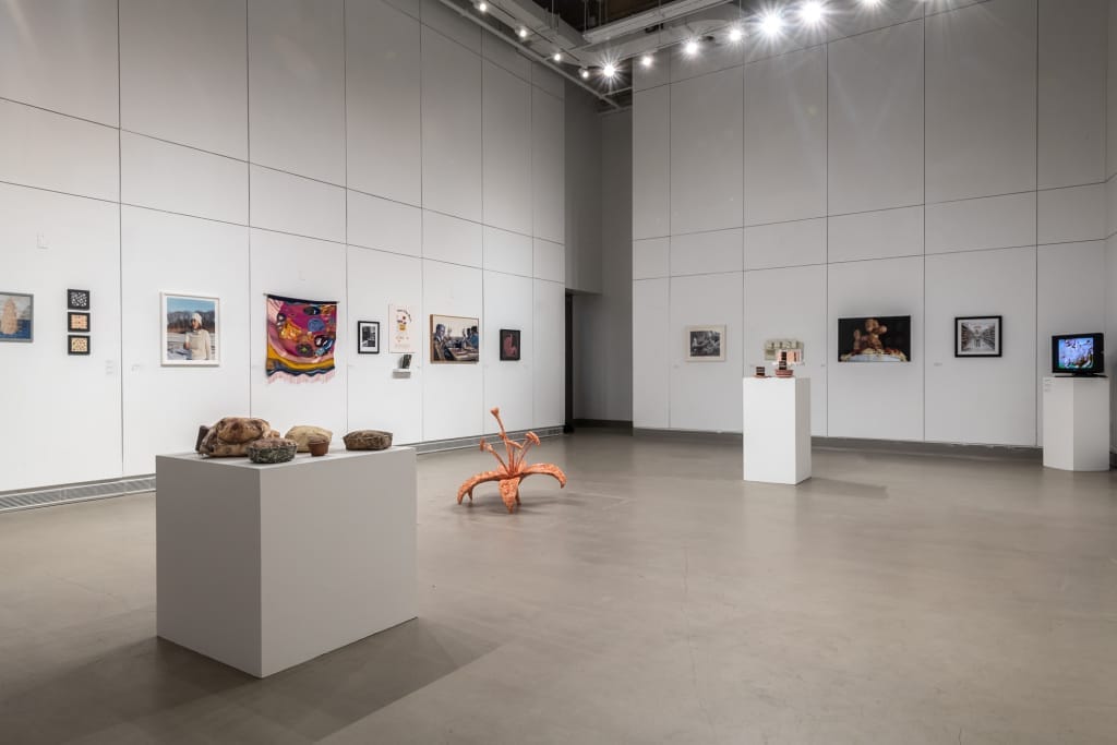 A spacious art gallery at MassArt Galleries, with high ceilings displaying various artworks, including framed paintings, a colorful tapestry, and sculptures on white pedestals. Spotlights illuminate the exhibits of contemporary art, enhancing the minimalist, modern atmosphere.