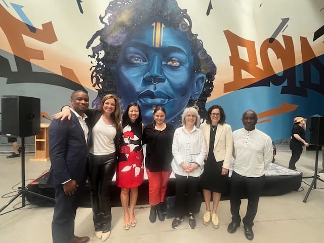 The Mayor of Boston, President of MasssArt and others stand in front of a Mural of a woman's face saying "Be Bold."