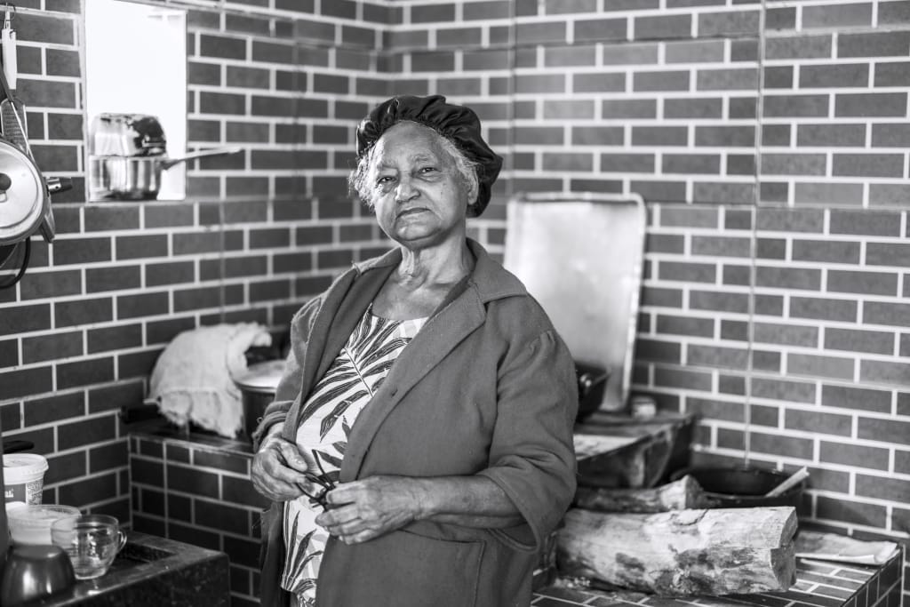 Elderly woman wearing a bonnet and patterned dress stands in a rustic kitchen with brick walls and wooden logs. She gazes thoughtfully, surrounded by various kitchen items. The scene is captured in black and white.
