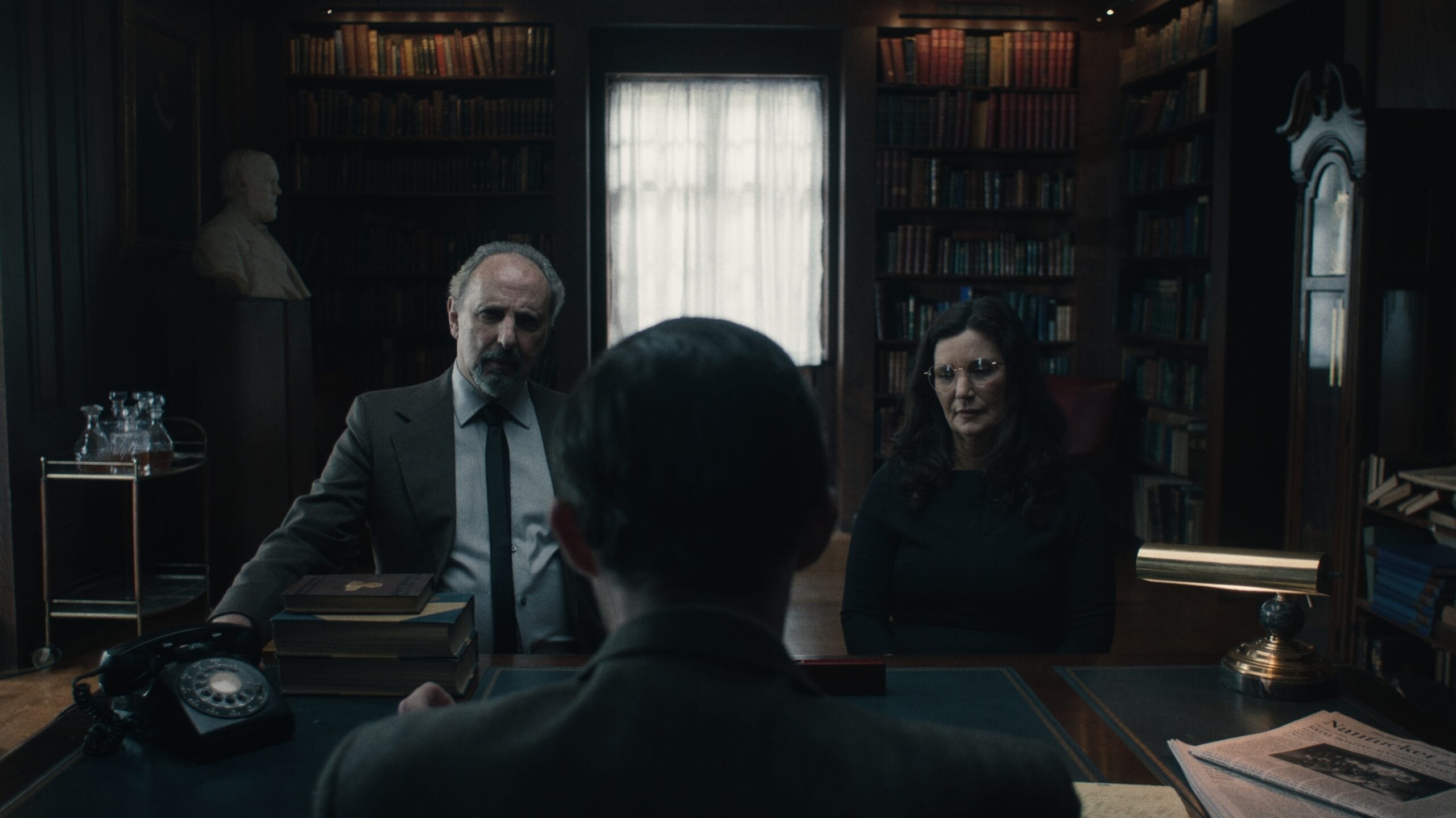 In a dimly lit office, a MassArt alum sits at a large desk covered in papers and books, facing two people. Shelves filled with books, a bust, and a vintage phone complete the scene evoking an atmosphere straight out of Dead Whisper, now available for streaming on demand.