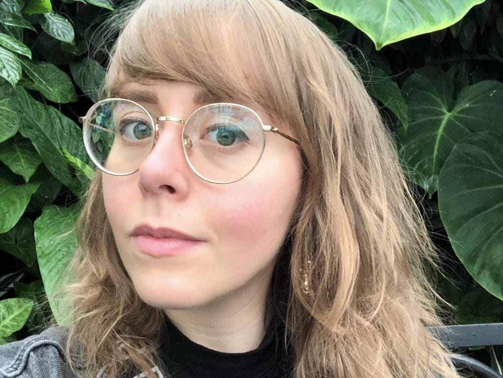 A person with long, wavy hair and glasses is looking at the camera. They are wearing a black top and standing in front of large green tropical leaves.