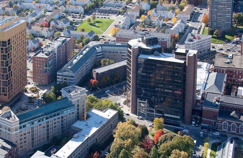 Aerial view of a cityscape showcasing the vibrant impact of autumn foliage mingling with residential and commercial buildings. In the foreground, a large modern building with reflective windows stands prominently, echoing the innovative spirit reminiscent of MassArts 2024 vision.