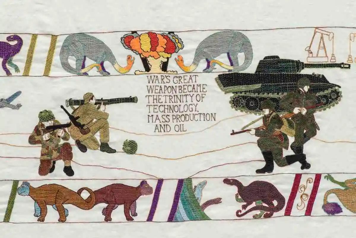 This exhibition panel features embroidered soldiers with weapons, a tank, and an explosion. The text reads, Wars Great Weapon Became the Trinity of Technology, Mass Production and Oil, with dinosaurs and an oil rig symbolizing the displacement caused by climate change.