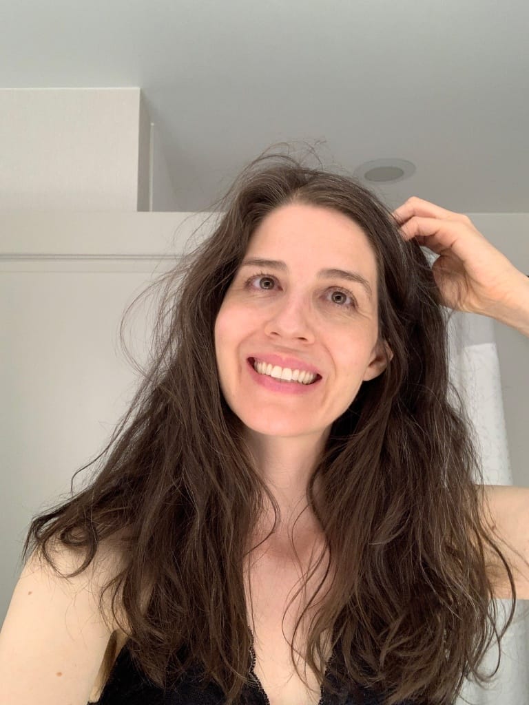 A person with long, dark hair smiles while standing indoors. They wear a black top and have one hand raised to their hair. The background is softly lit with a doorway visible.