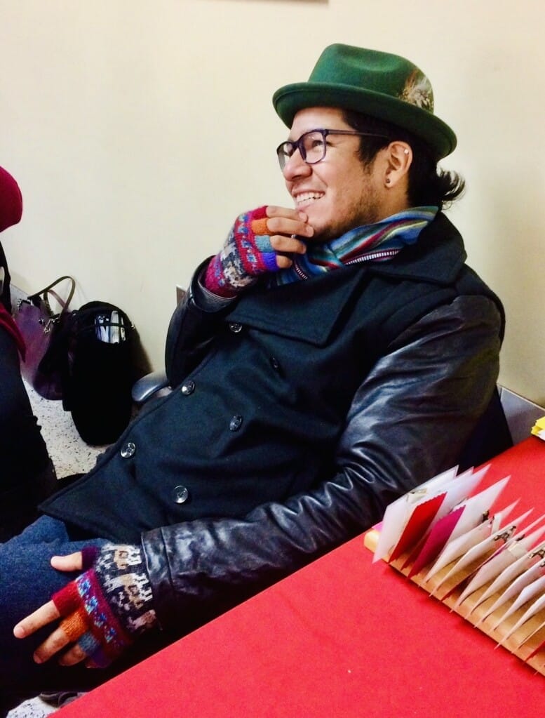 A person wearing a green hat, glasses, and colorful gloves smiles while sitting at a table with red and white folders. They are dressed in a black coat with leather sleeves and a colorful scarf. A black bag is visible in the background.