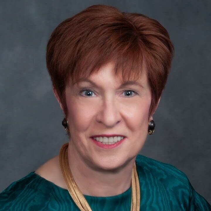A person with short auburn hair, wearing a gold necklace and an emerald green top, smiles confidently at the camera against a gray background, embodying the professional grace of a member of the Board of Trustees.
