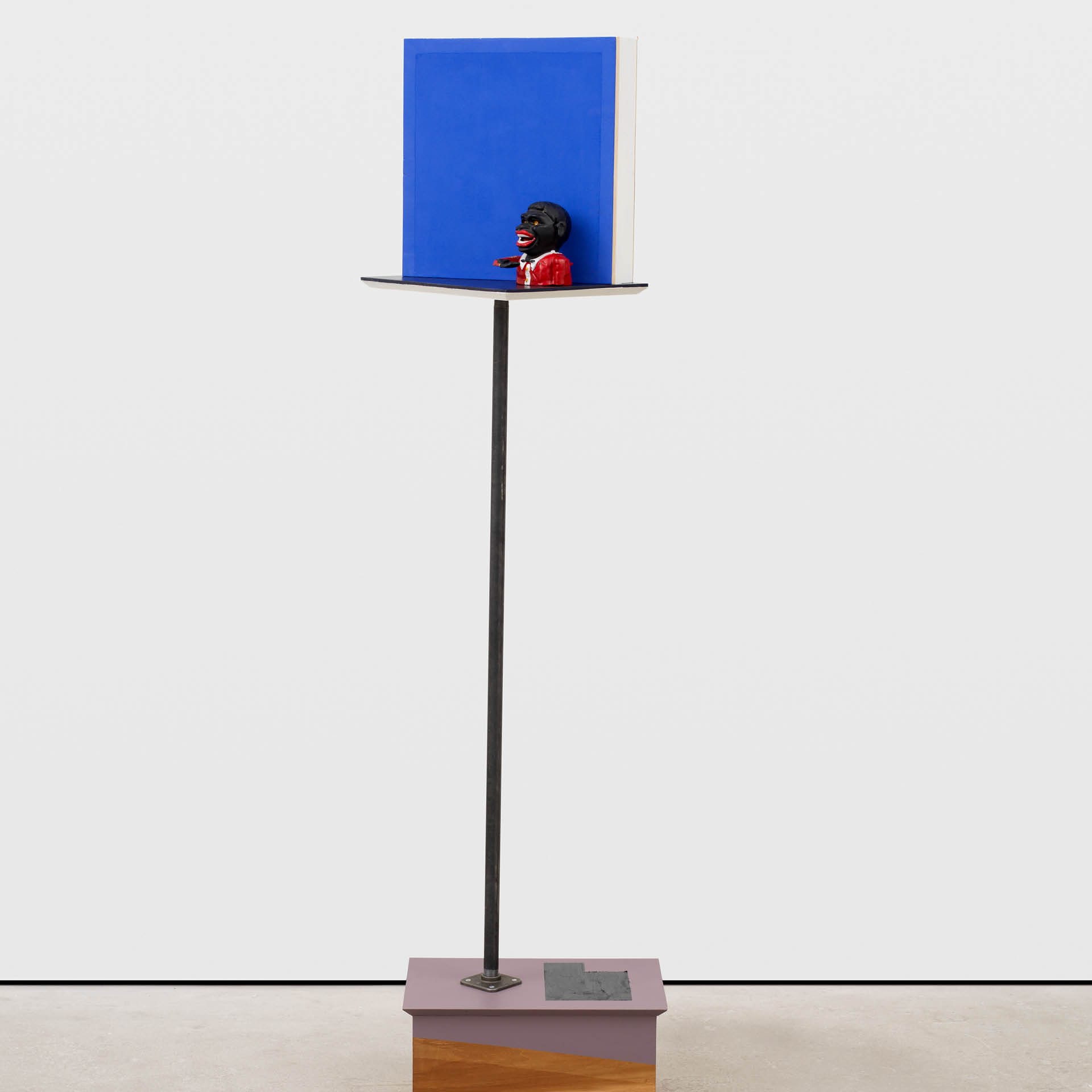 A modern art sculpture featuring a small figurine with dark skin and a red shirt holding a book stands on a high pedestal. Behind it is a tall blue rectangle. The base is a simple wooden block. The background is plain white.