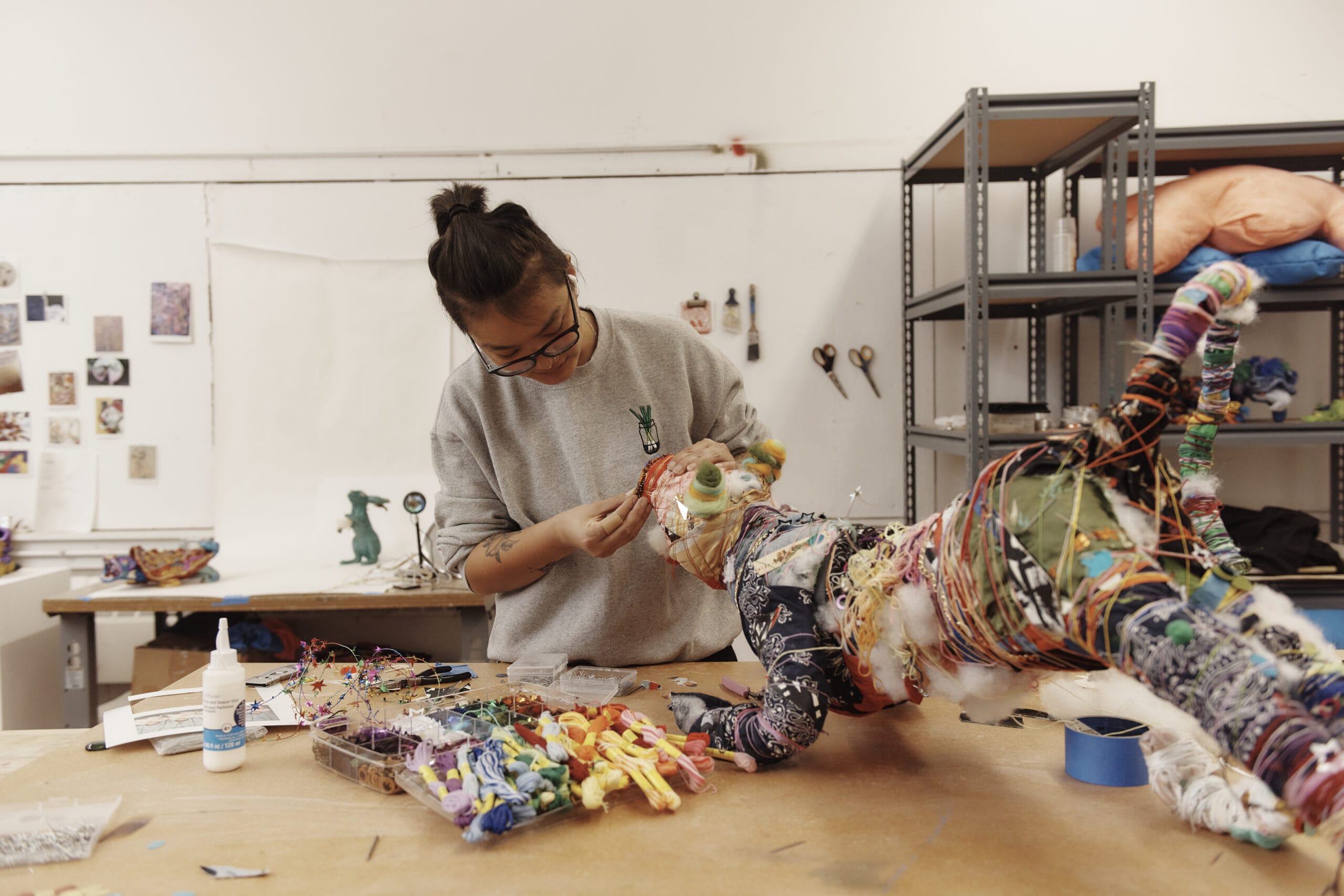 A person with a Studio Art Certificate is working on a colorful, fabric-covered sculpture in an art studio. They focus intently on the details, surrounded by art supplies and tools. The bright space features white walls and shelves in the background.