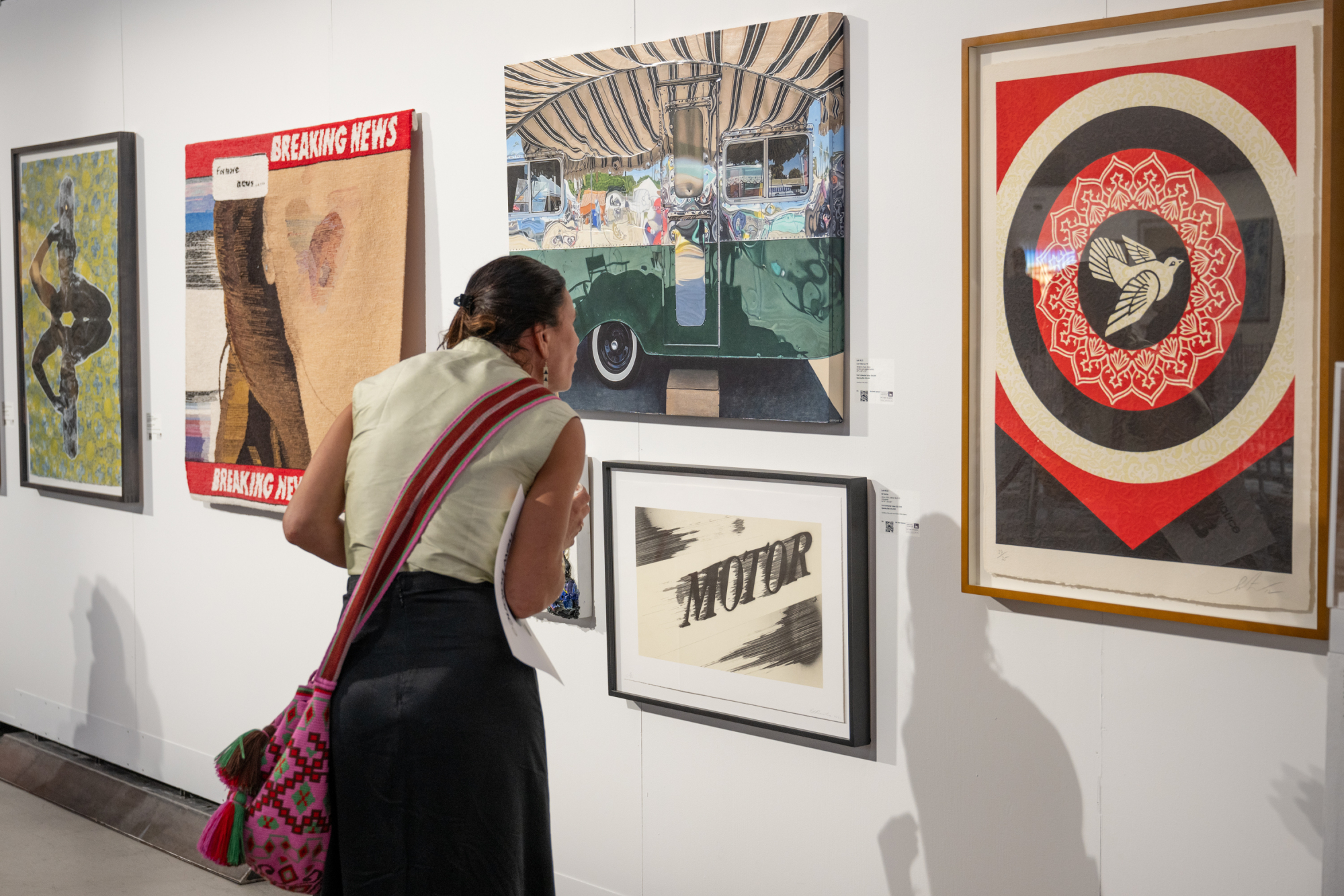 A person leans in to closely examine artwork displayed on a gallery wall. Among the colorful pieces, sponsored by esteemed partners, a framed image of a bird in a circle captures their attention. The person has a colorful bag and wears a sleeveless top.