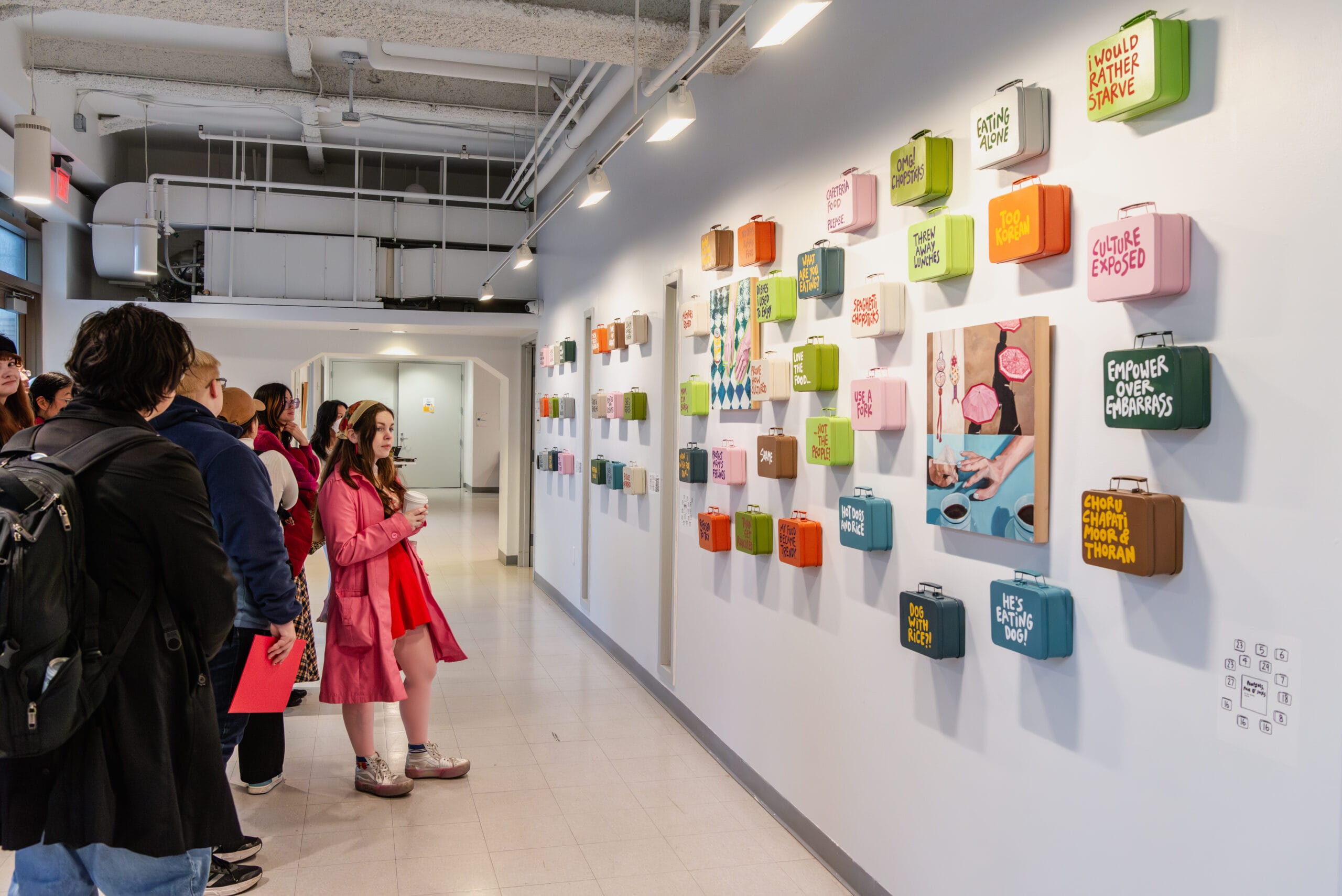 A class field trip of students crowd into a gallery to observe colorfully painted lunch boxes hung across a wall.