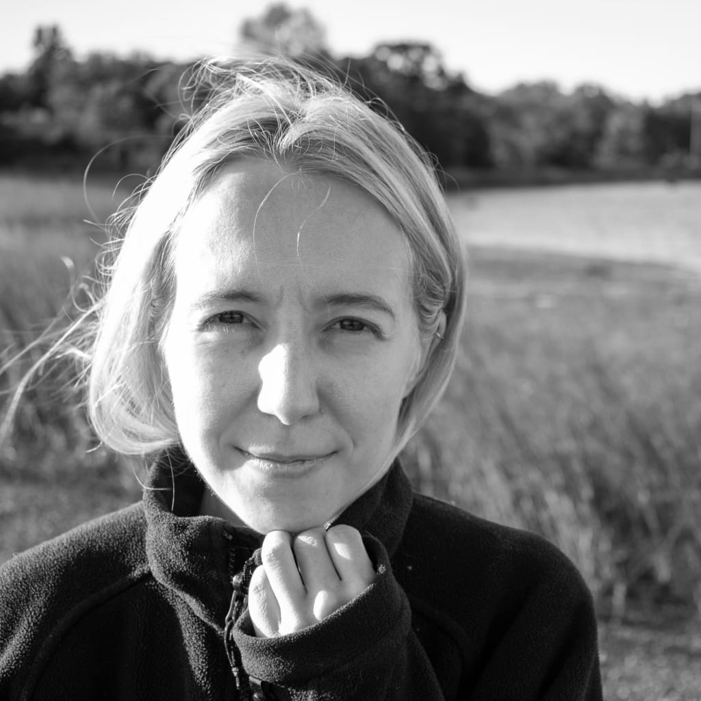 A person with short hair is outdoors, wearing a zip-up jacket, and gently holding the collar. The background features a blurred landscape with grass and water, suggesting a natural setting. The image is in black and white.