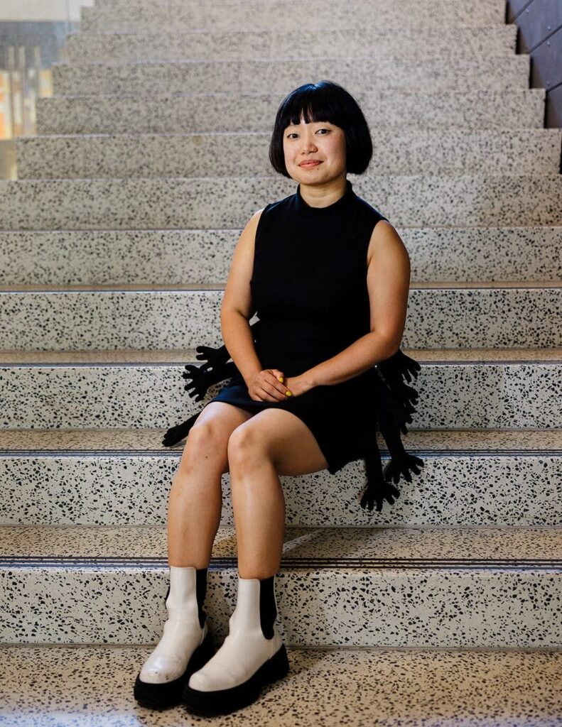 Caroline Hu sits on a staircase wearing a black dress and white boots.