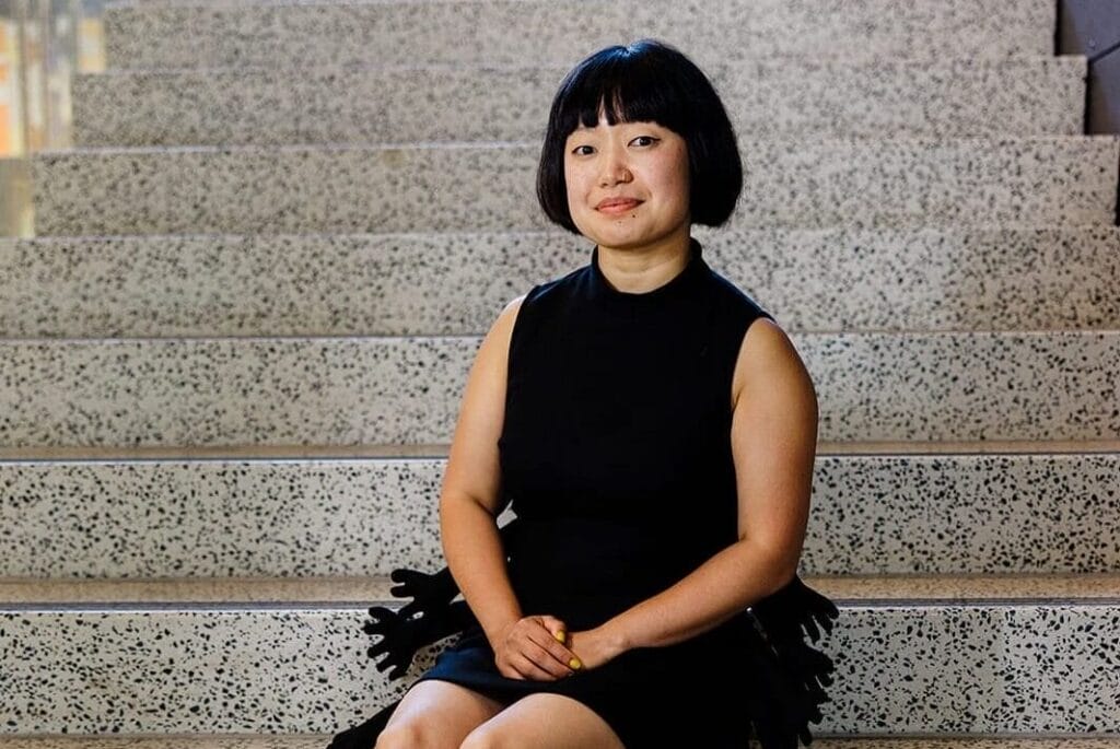 Caroline Hu sits on a staircase wearing a black dress and white boots.