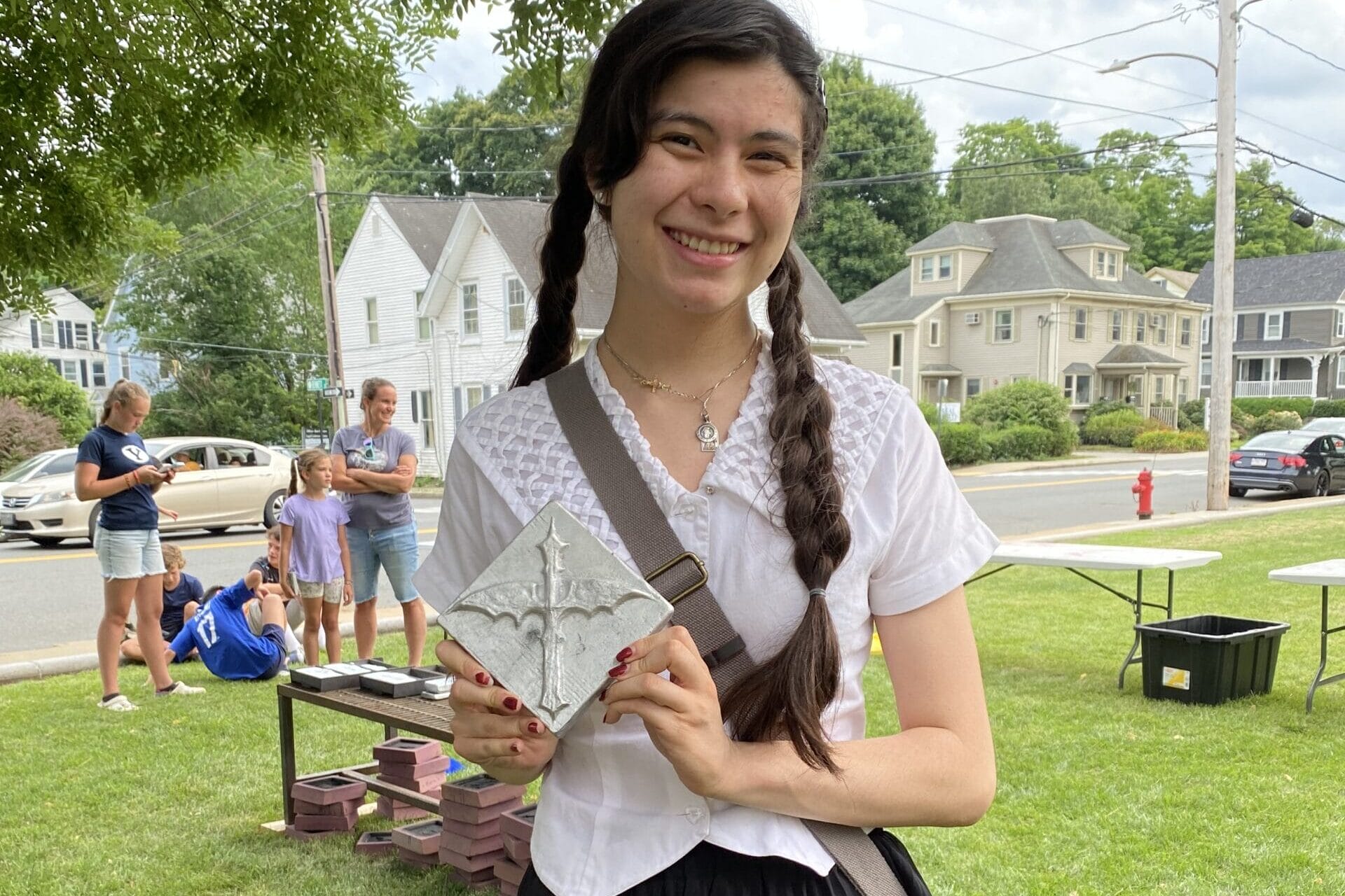 A young woman with braided hair stands on grass, holding a decorative concrete tile that showcases her creative process. She wears a white blouse and black skirt, smiling under a tree. People and houses are in the background, reminiscent of a scene near MassArts vibrant campus.