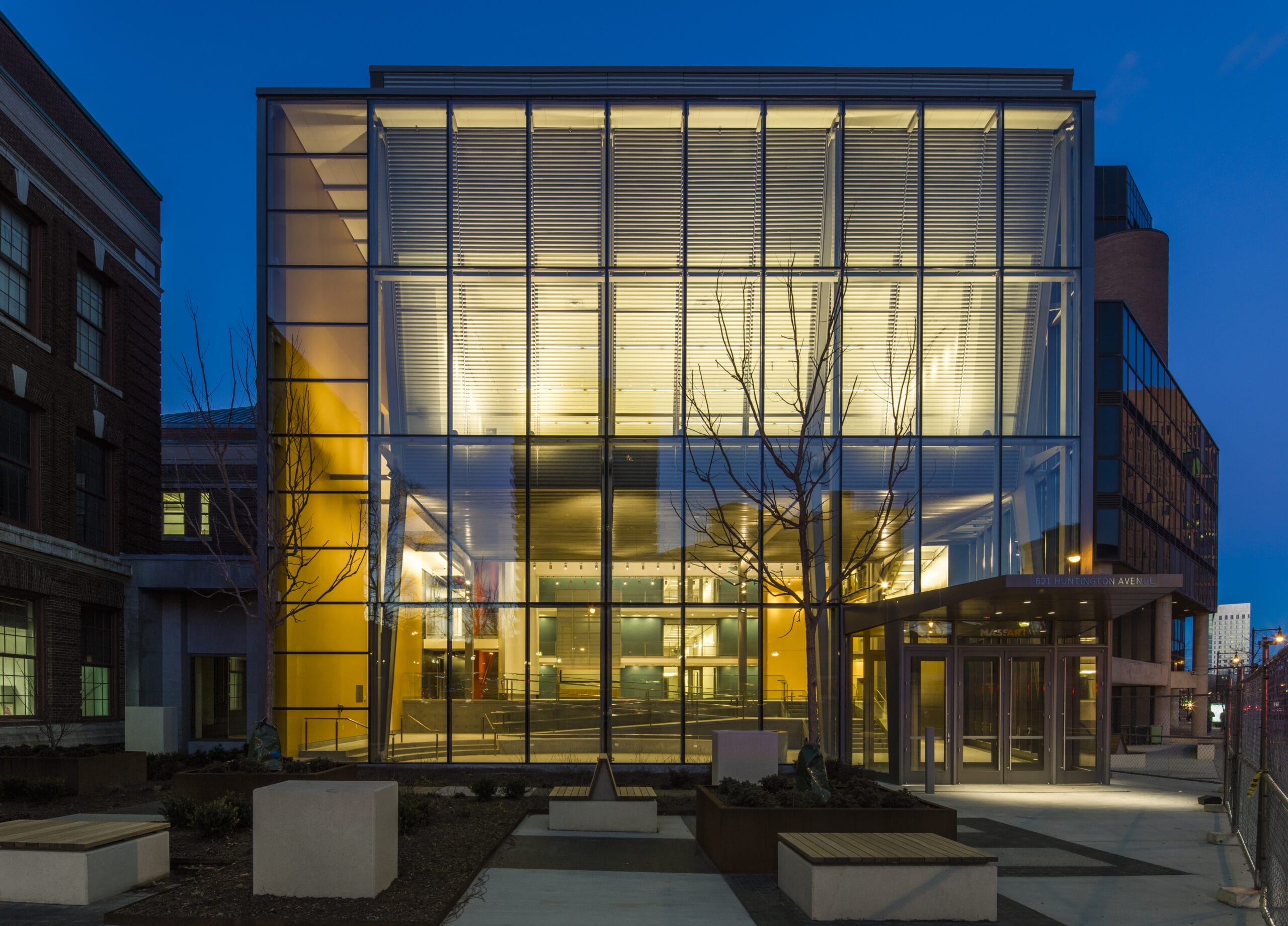 A modern building with a glass facade illuminated at night, perfect for conferences. The structure showcases a spacious, well-lit interior with clean lines and metal accents. Benches and a paved walkway are visible in the foreground, ideal for hosting event services.