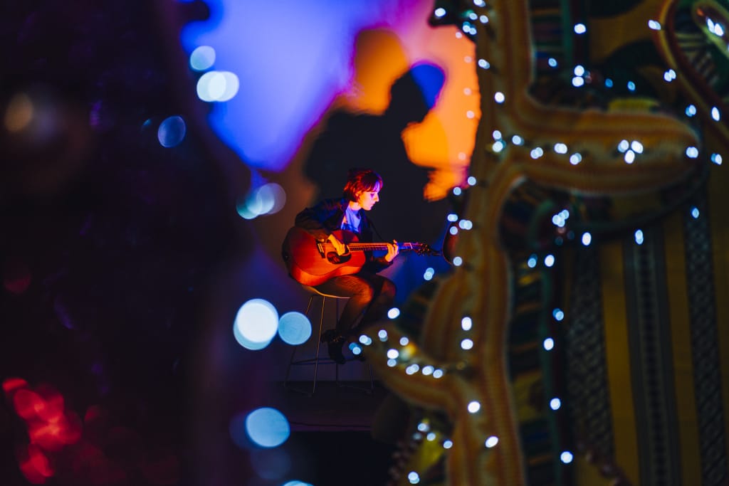 A woman plays guitar in a colorfully lit performance space.