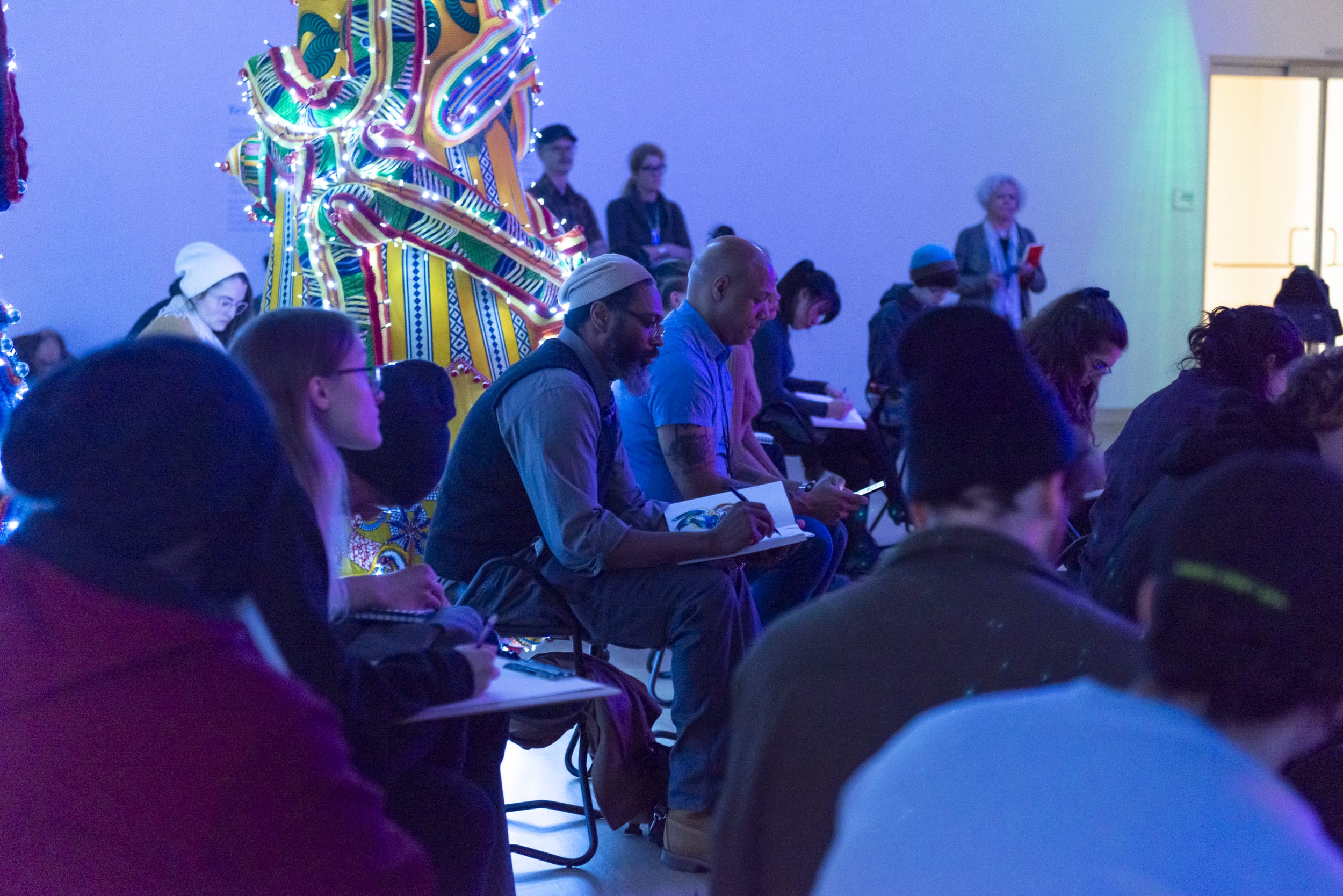 A large gallery room full of people sitting and drawing in a blue light tinted room.