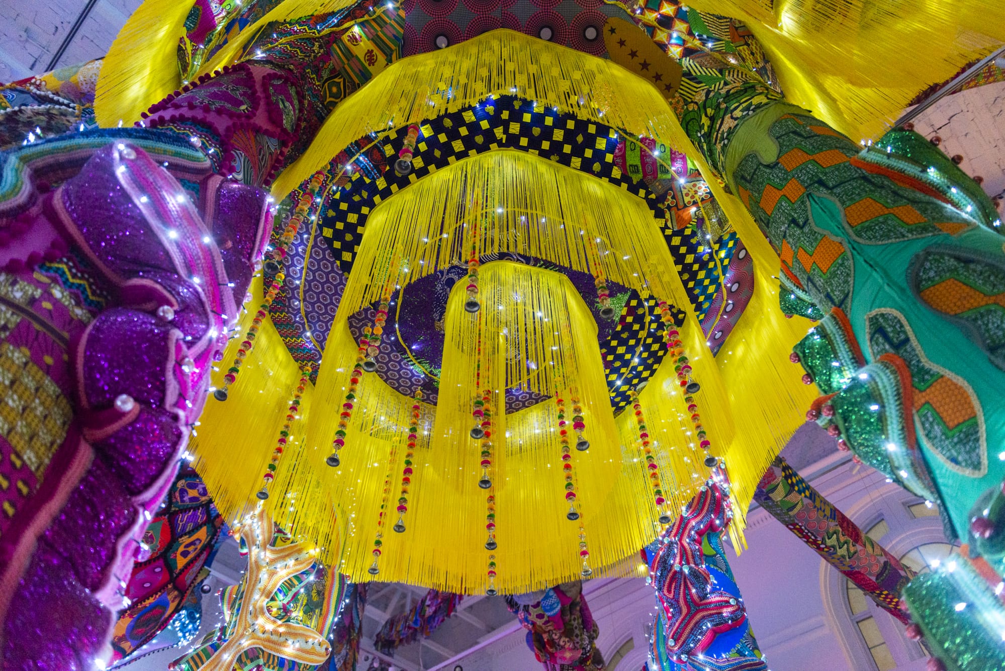 Yellow strands of fiber hang down from the base of a colorful, sculptural, fabric artwork installation.