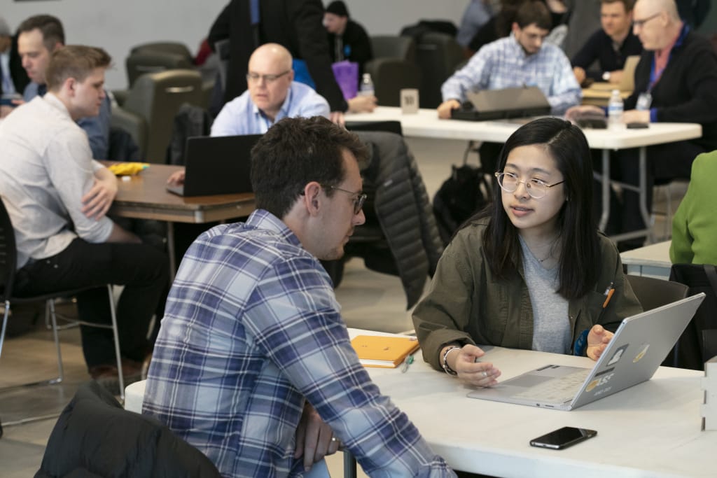 Students sit at tables with their laptops, talking to professionals.