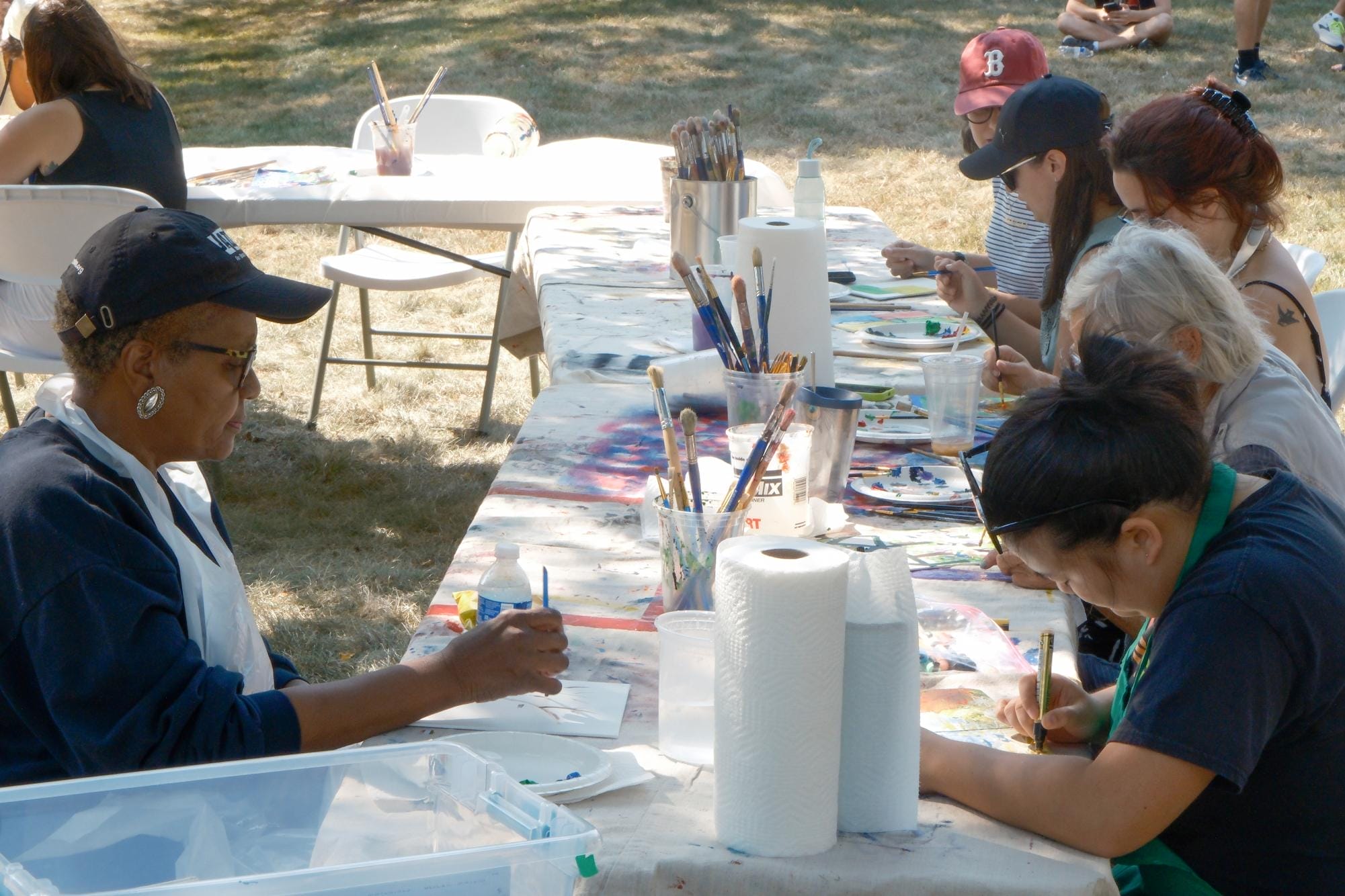 People sitting around long white tables in dappled sunlight with with paints and paint brushes.