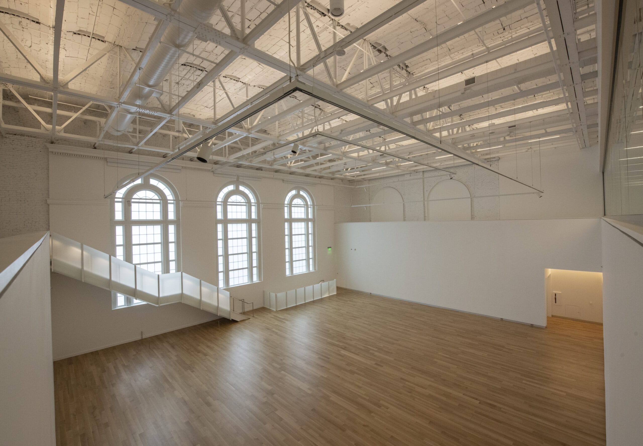 Spacious, empty gallery with high ceilings, exposed beams, and large arched windows letting in natural light. Perfect for conferences or event services, the wooden floor and white walls create a minimalist aesthetic, while a staircase with illuminated steps adds a modern touch.