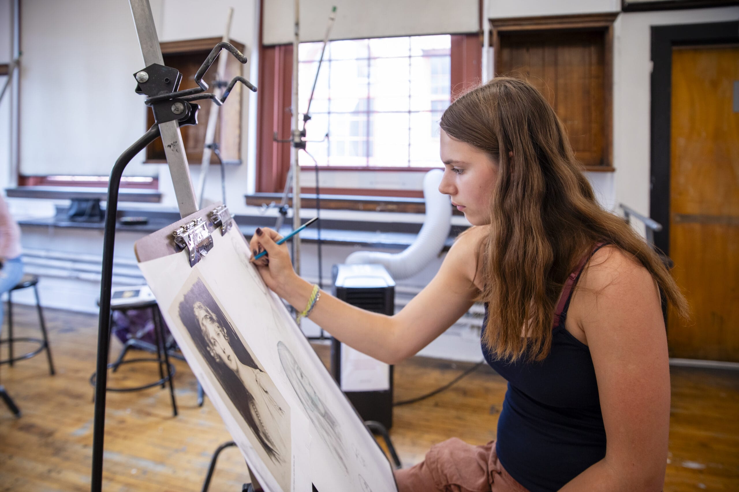 A high school student sits at an easel drawing in a brightly lit room.