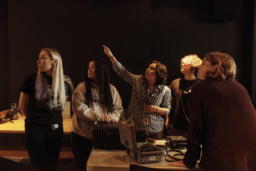 A group of five people in a dimly lit room stands around a table with electronic equipment. One person is pointing upwards, while others look in the same direction with attentive expressions, illustrating their shared commitment to career development and an inquisitive approach to progress.