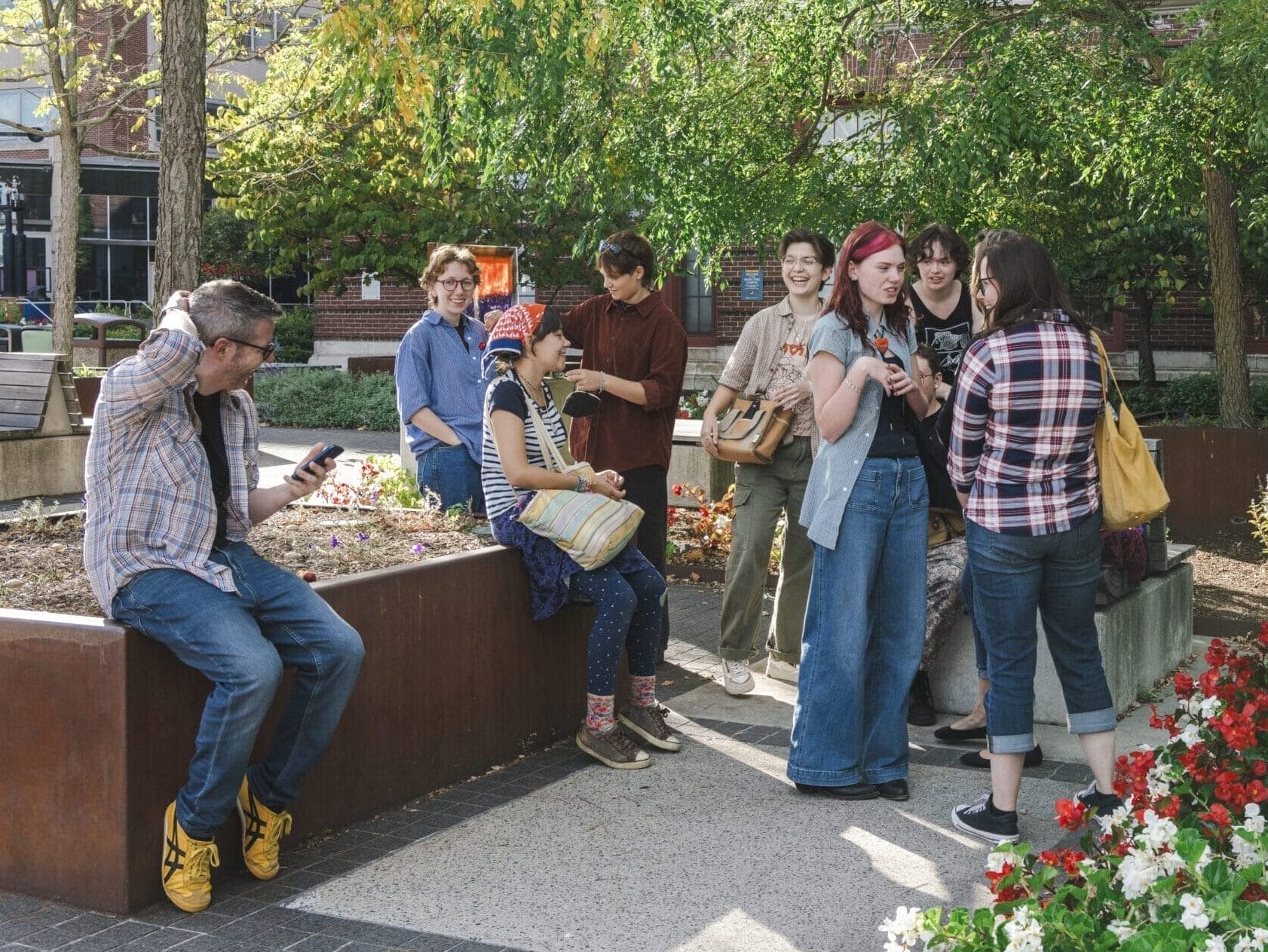 A group of people gather outdoors in a garden area, some sitting and others standing while one person checks their schedule on a phone. They are surrounded by trees and flowers, with a brick building in the background. The scene is casual and social.