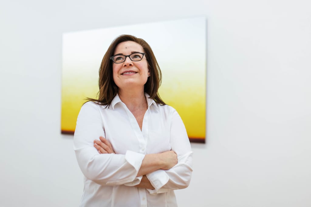 Headshot of Nicole Chesney with her arms crossed in front of a white and yellow painting on the wall.