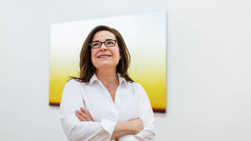 Headshot of Nicole Chesney with her arms crossed in front of a white and yellow painting on the wall.