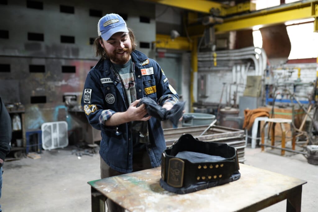 A person in a workshop smiles while folding a piece of dark fabric. They wear a blue jacket with various patches and a blue cap. A workbench with a leather apron or tool belt is in front of them. The background shows industrial equipment and walls.