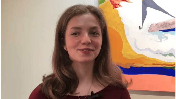 A person with long brown hair, likely a Creative Director, smiles while standing in front of a colorful abstract painting. Wearing a burgundy top, they have a microphone clipped to their clothing, blending their passion for art and civic engagement.