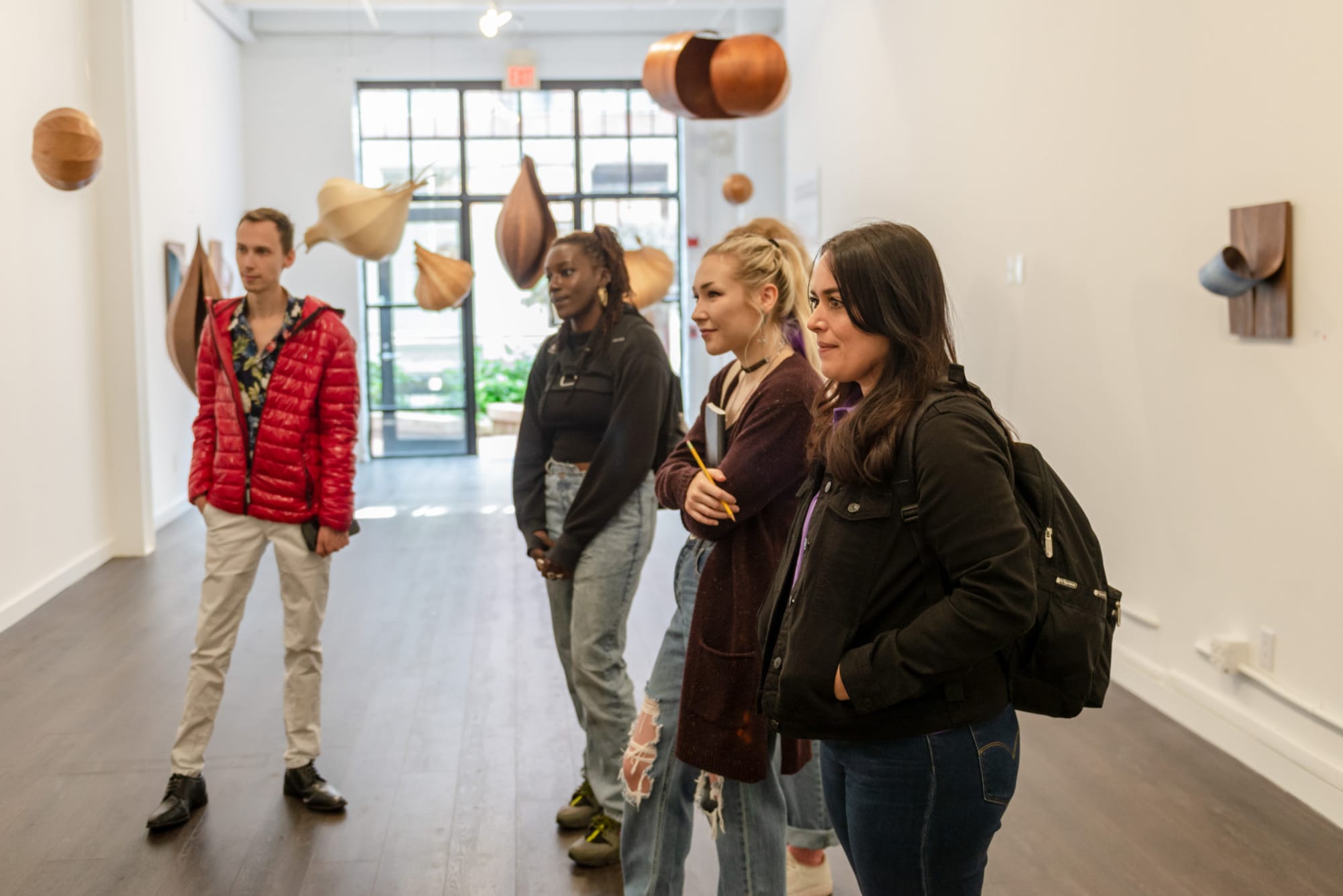 Four people stand in an art gallery, visiting a captivating display of wooden sculptures hanging from the ceiling. The room is well-lit with natural light from large windows, enhancing the varied shapes and sizes of the artwork.