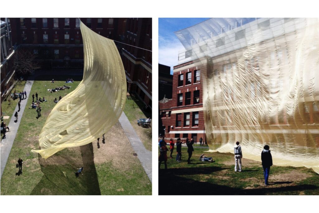 Two photos show Gold Divide, a large, semi-transparent yellow fabric sculpture billowing between red brick buildings in a grassy courtyard as people walk, sit, and observe the installation from different angles.