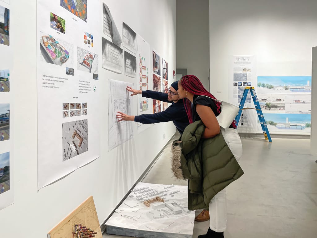 Two graduate students pin up architectural drawings in a gallery.