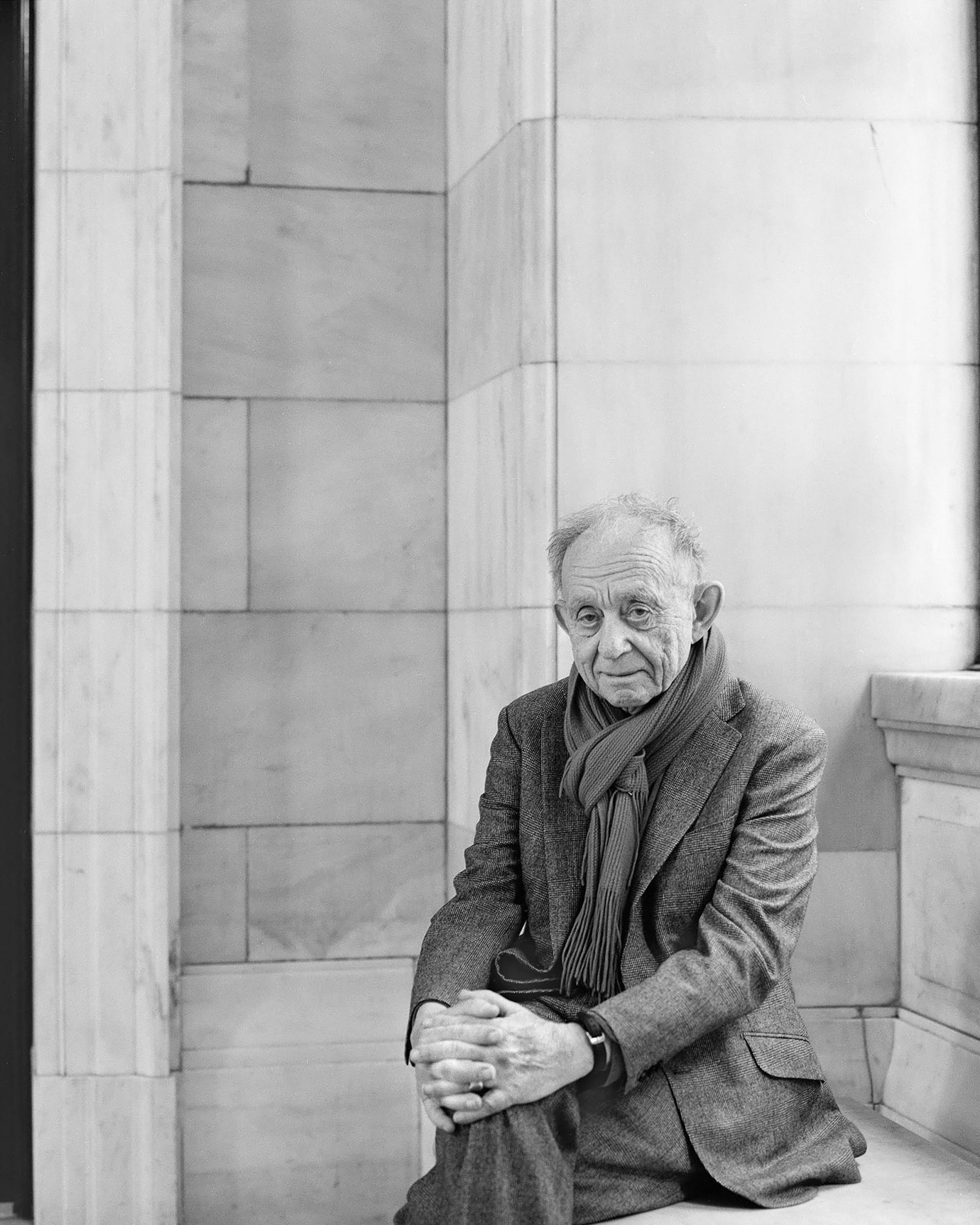 An elderly man in a suit and scarf sits on a bench, hands clasped, against a backdrop of marble walls. The black-and-white image conveys a sense of contemplation, reminiscent of the honor found at the Common Good Awards at MassArt.