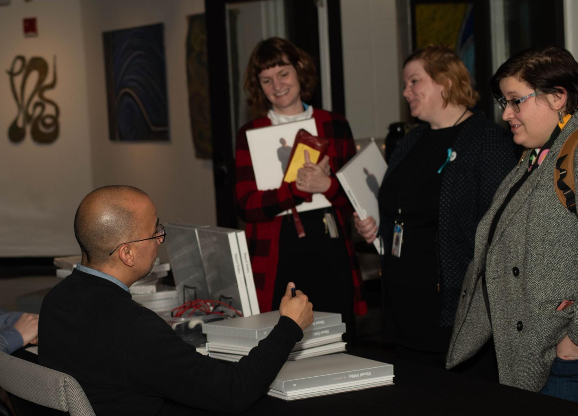 A person with glasses is seated at a table, signing books for three people. The three individuals are holding books and papers, engaging in conversation and smiling. Artworks and framed pictures are visible on the walls in the background.