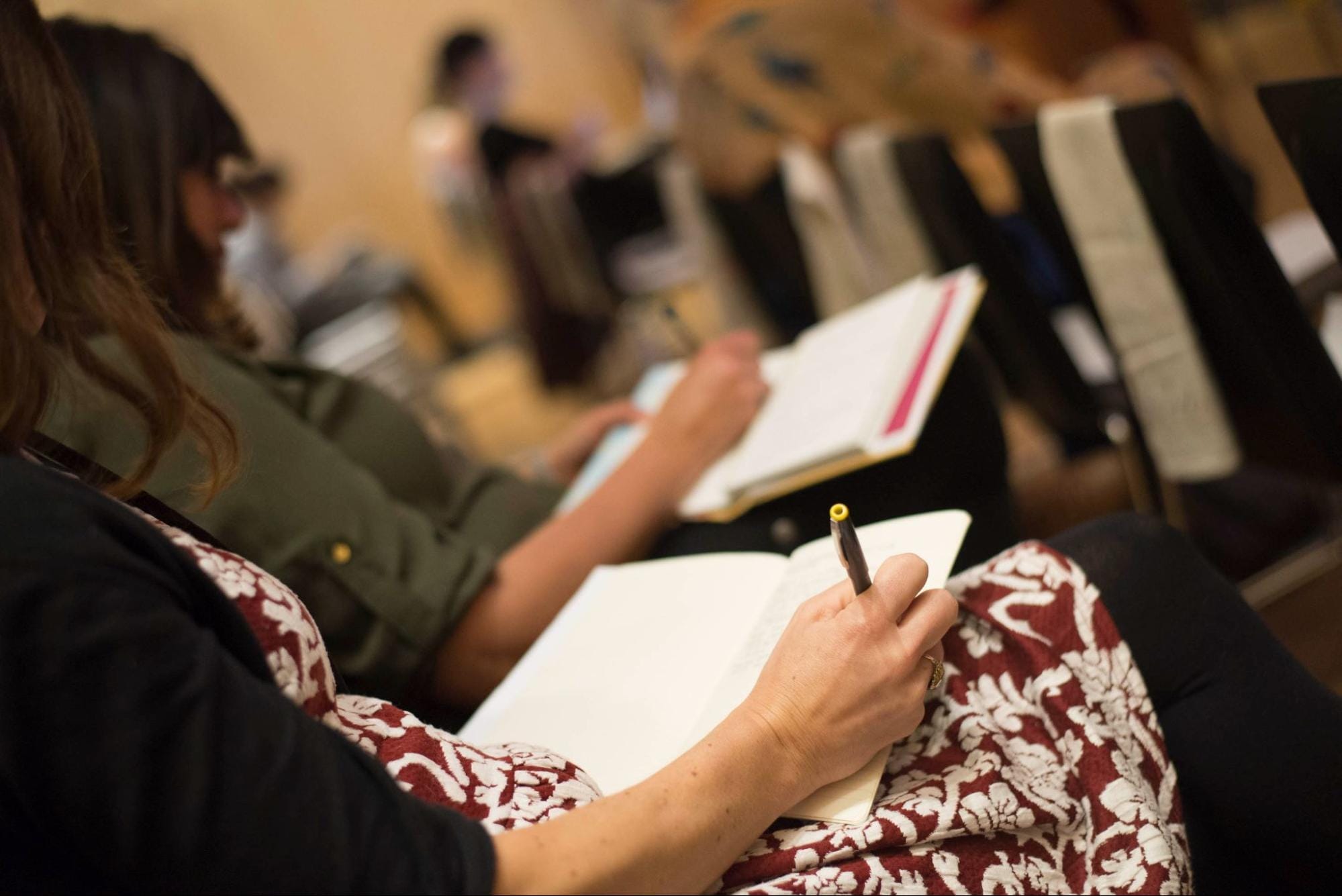 People sitting in a row taking notes in notebooks during a meeting or lecture. The foreground shows a person wearing a patterned outfit holding a pen. The background is softly blurred, focusing on participants attentive engagement.