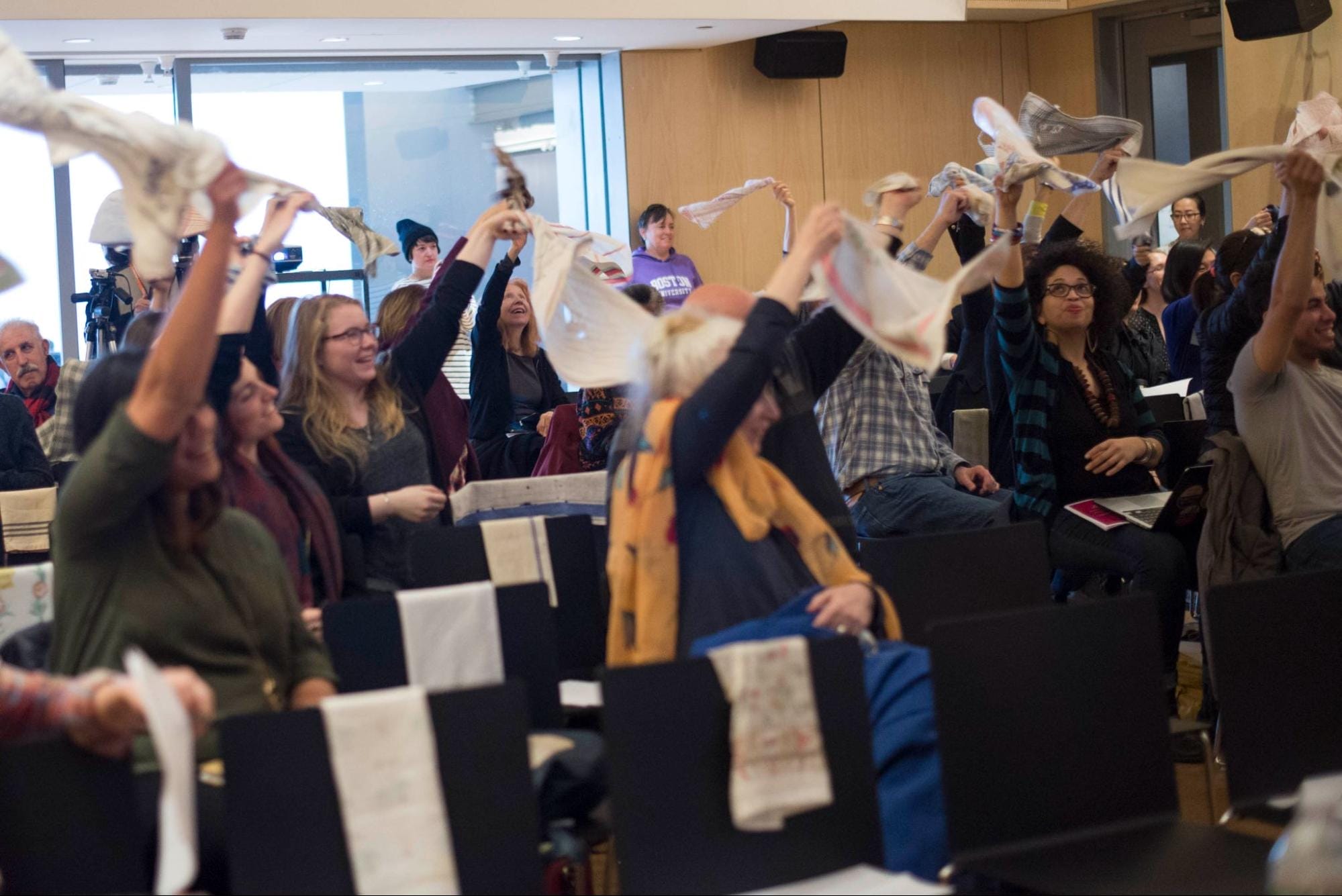 A group of people in a lecture hall wave white handkerchiefs in the air, looking engaged and enthusiastic. The room has rows of black chairs and a large window in the background.