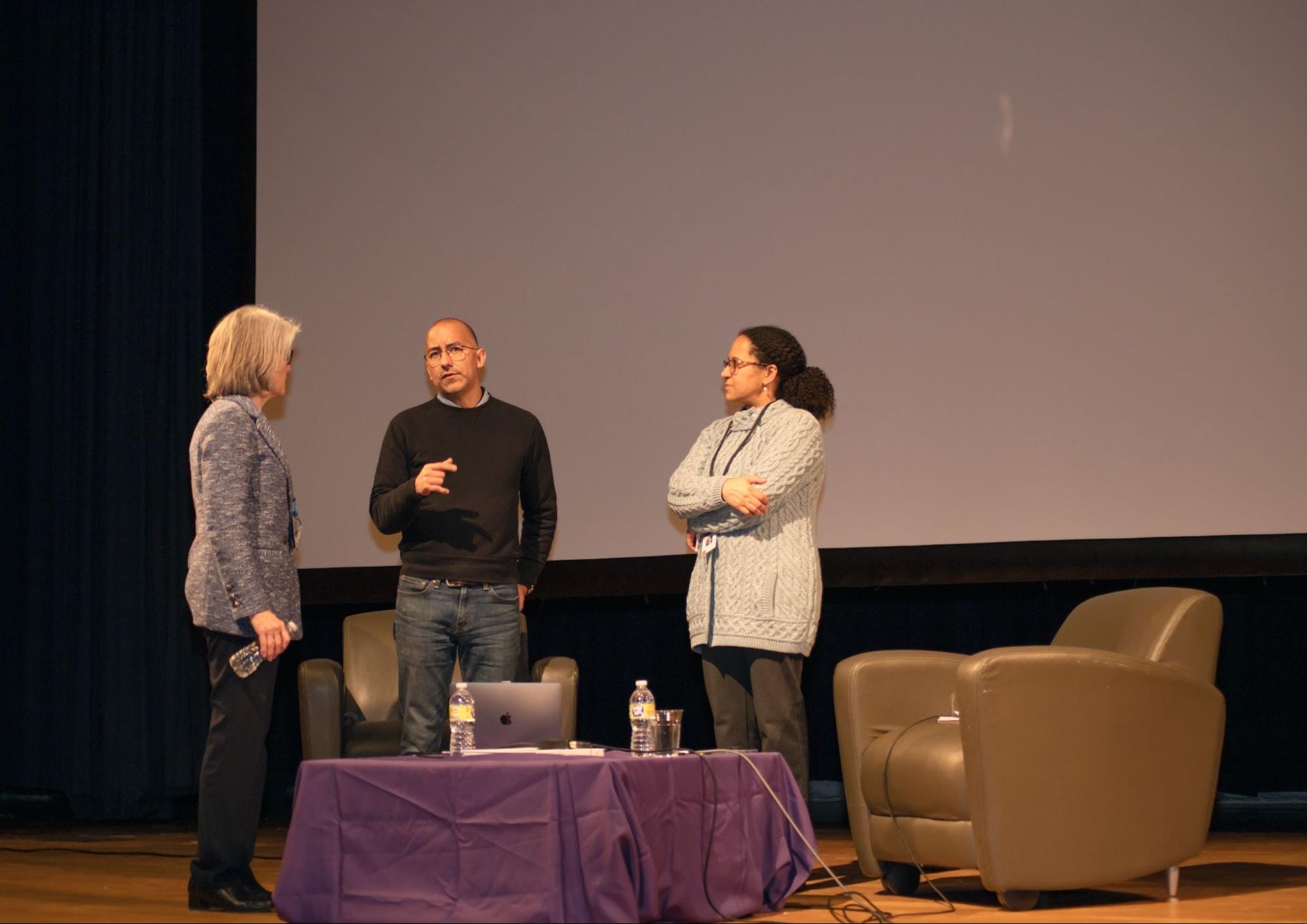 Three people stand on a stage in front of a large screen, engaging in conversation. Two armchairs are visible on the right, and a table with a laptop and water bottles is in front of them. The setting appears to be a lecture or presentation.