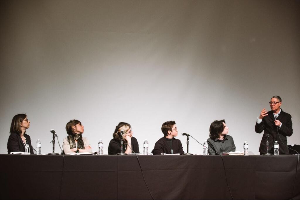 A panel of six people sitting at a long table with microphones. Five are seated, listening attentively, while one, standing, gestures as they speak. Bottles of water are placed on the table in front of each person.