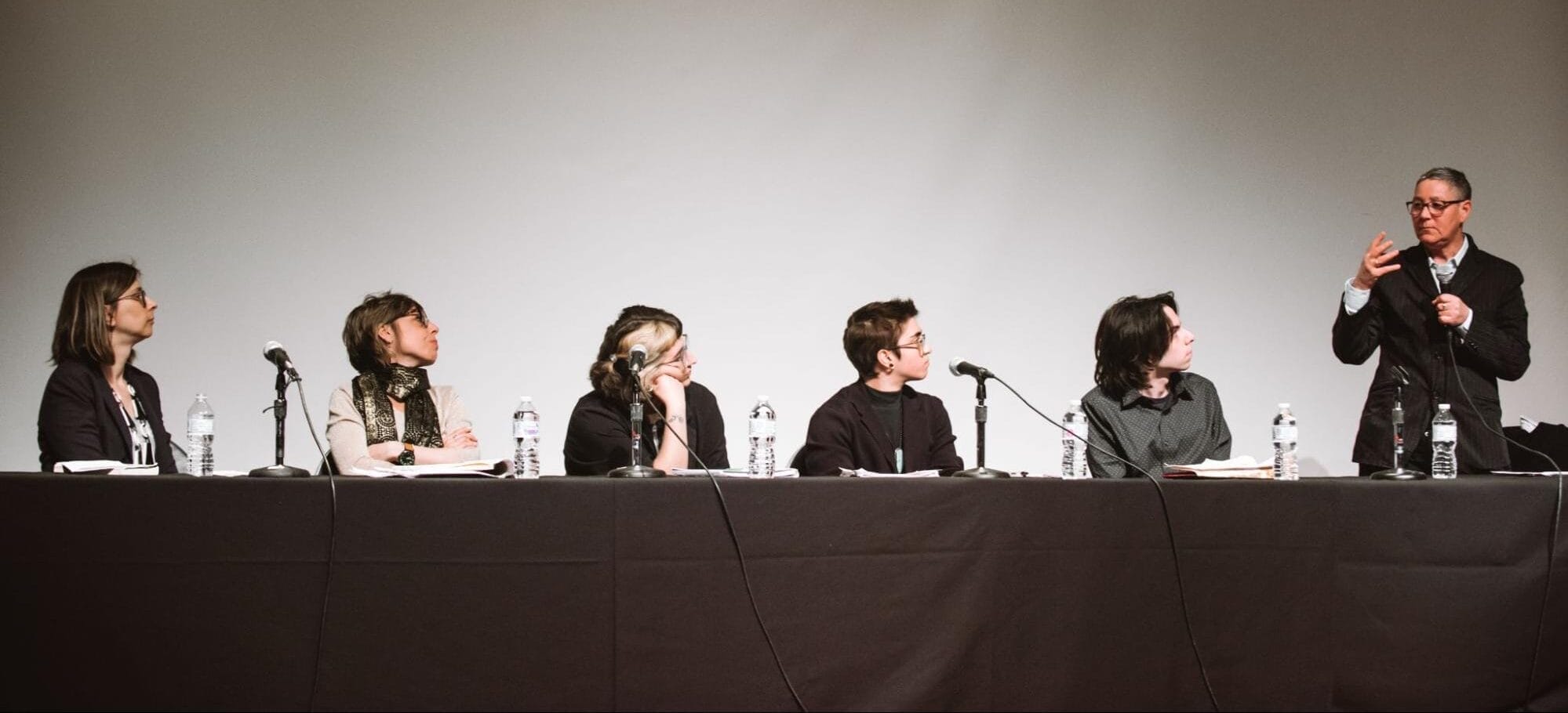 A panel of six people sitting at a long table with microphones. Five are seated, listening attentively, while one, standing, gestures as they speak. Bottles of water are placed on the table in front of each person.