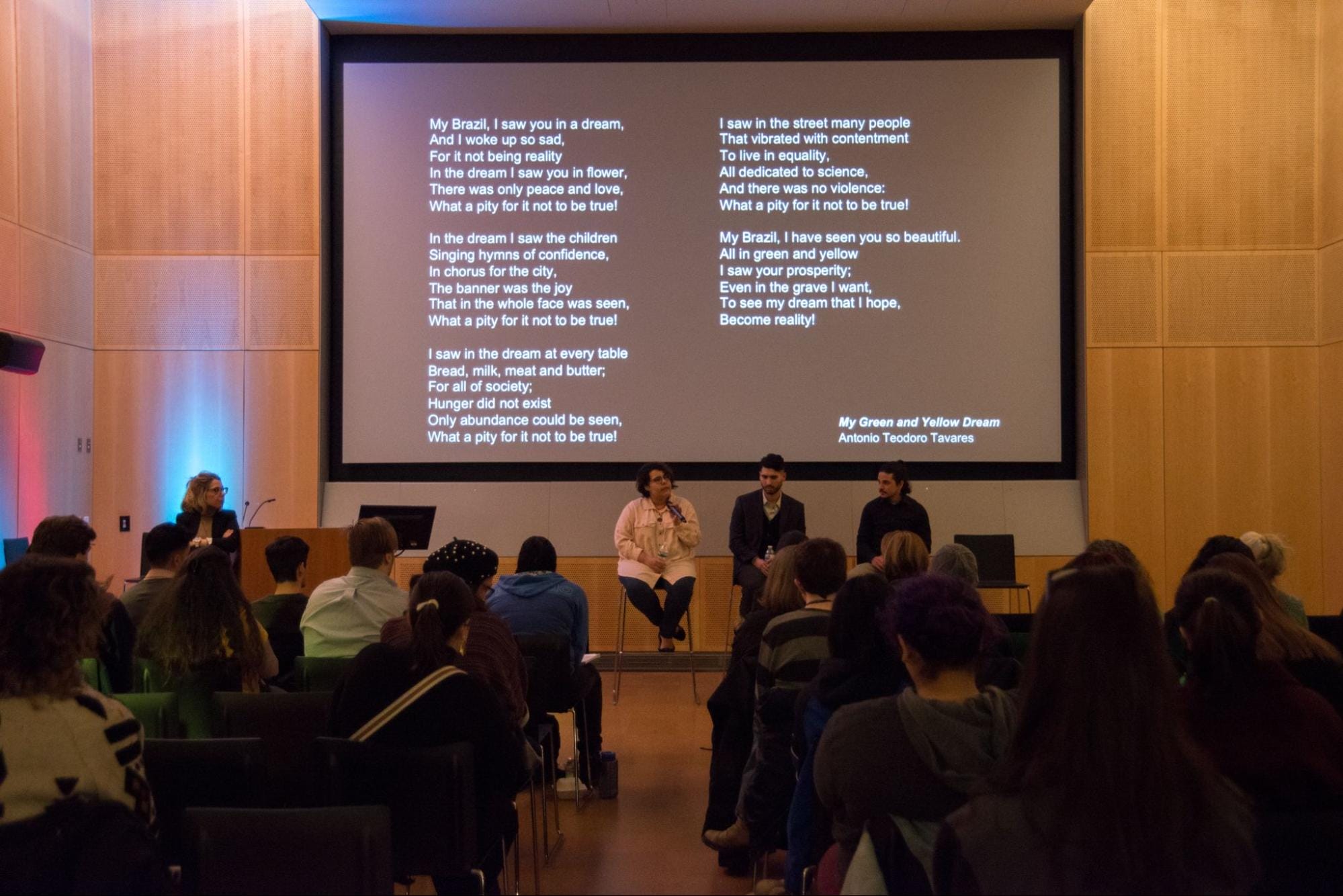 A panel of four people sit on stage in front of an audience. A large screen behind them displays a poem titled My Brazil by Antonio Teodoro Travers, depicting dreams and disappointments. The audience is seated in a dimly lit room.